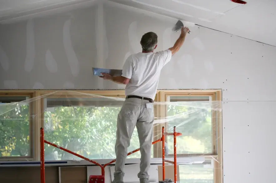 Drywall finisher taping a vaulted ceiling with the windows sealed off by plastic sheeting